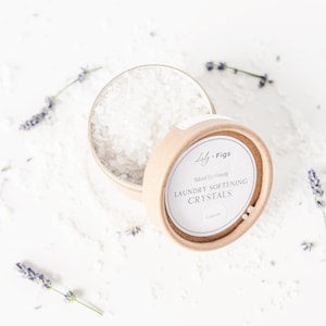 Può includere: Un contenitore rotondo di cartone, parzialmente aperto, pieno di cristalli ammorbidenti bianchi per bucato. L'etichetta recita "Lily + Figs, Laundry Softening Crystals, Lavender". Rametti di lavanda sono sparsi intorno al contenitore.