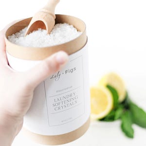 Op de afbeelding: Een witte en bruine container met wasverzachtende kristallen en een houten schep. Het etiket luidt "Lily + Figs" en "Laundry Softening Crystals". Het nettogewicht is 1,13 kg. Citroenen en muntbladeren zijn op de achtergrond.