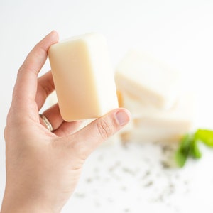 Op de afbeelding: Een hand die een lichtgekleurd, rechthoekig stuk zeep vasthoudt. Verschillende andere stukken zijn enigszins onscherp op de achtergrond. De zeep lijkt ongeparfumeerd en natuurlijk te zijn. De achtergrond is wit.