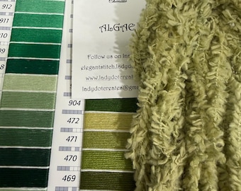 May include: A skein of light green hand-dyed eyelash chenille yarn labeled Algae, 100% cotton, 9 yards. It is positioned next to a DMC color chart featuring various shades of green embroidery floss with numbered labels for color identification.
