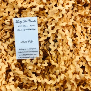 May include: A close-up of a pile of gold-colored rickrack trim. The trim is made of 100% cotton and is 3 yards long. The card reads "Lady Dot Creates" and "Gold Fish".