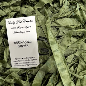 May include: A pile of olive green hand-dyed rayon ribbon with a white product label that reads Lady Dot Creates, 100% Rayon - 3 yards, Hand-Dyed ribbon, DRUM ROLL GREEN, and contact information for Instagram, a website, and email.