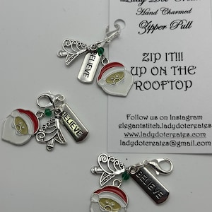 May include: Three silver-toned metal zipper pulls featuring a Santa Claus face charm with a red hat, a filigree tree charm, and a rectangular tag engraved with the word BELIEVE, all attached to a lobster clasp with a small green bead.