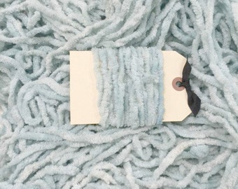 May include: A close-up of a light blue fuzzy yarn with a white tag attached. The tag has a black ribbon tied around it.