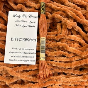 May include: A skein of hand-dyed chenille yarn in a burnt orange color called "Bittersweet". The yarn is 100% cotton and is 3 yards long. The label also includes the brand name "Lady Dot Create" and contact information.