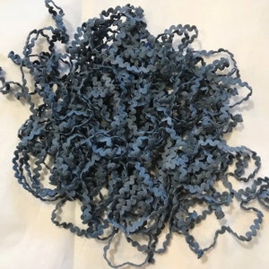 May include: A pile of blue-gray ric rac trim with a wavy, zig-zag edge. The trim is a mix of light and dark shades of blue, creating a textured appearance. This decorative trim is suitable for sewing and craft projects.