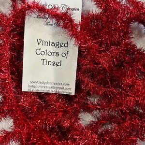 May include: Red tinsel garland, a vintage Christmas decoration. The garland is made of shiny, metallic strands and is perfect for adding a touch of sparkle to your holiday decor. The image also includes a business card with the text "Vintaged Colors of Tinsel" and contact information for "Lady Dot Creates".