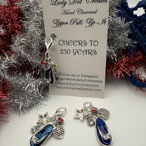 May include: Two hand-charmed zipper pulls with a patriotic theme. Each charm features a blue and silver flip-flop, a star, a heart with an American flag design, and a red gem. The card reads "CHEERS TO 250 YEARS."
