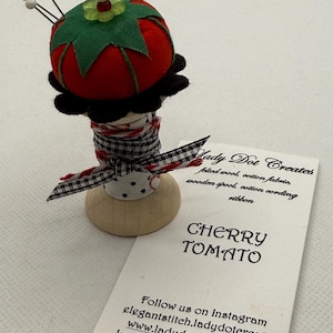 May include: A handmade cherry tomato pincushion. The red felt tomato has green leaves and is mounted on a wooden spool. The spool is wrapped with fabric and ribbon. The tag reads "CHERRY TOMATO".