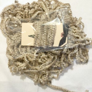 May include: A close-up of a bag of beige and brown pom-pom trim. The trim is in a clear plastic bag with a white tag that says "The Ribbon Retreat".