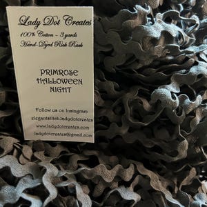 May include: A close-up of a pile of dark gray rickrack trim. The trim is 100% cotton and is hand-dyed. The label reads "Lady Dot Creates", "100% Cotton - 3 yards", "Hand-Dyed Rick Rack", "Primrose Halloween Night", "Follow us on Instagram", "elegantstitch.ladydotcreates", "www.ladydotcreates.com", "ladydotcreates@gmail.com".