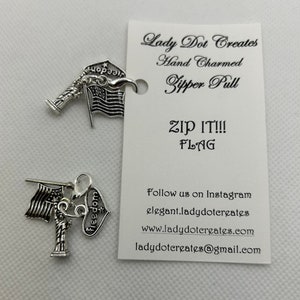 May include: Two silver zipper pulls featuring an American flag design. The top pull has a small sign that says "Hope" and the bottom pull has a heart that says "Freedom". Both pulls have a small silver replica of the Statue of Liberty. The card behind the pulls says "Lady Dot Creates Hand Charmed Zipper Pull ZIP IT!!! FLAG Follow us on Instagram elegant.ladydotergates www.ladydotergates.com ladydotereates@gmail.com"