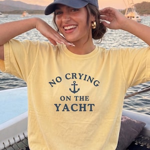 May include: Yellow t-shirt with the text "NO CRYING ON THE YACHT" in navy blue, featuring an anchor graphic. The person in the image is wearing a navy blue baseball cap and gold hoop earrings. The background shows a body of water and boats.