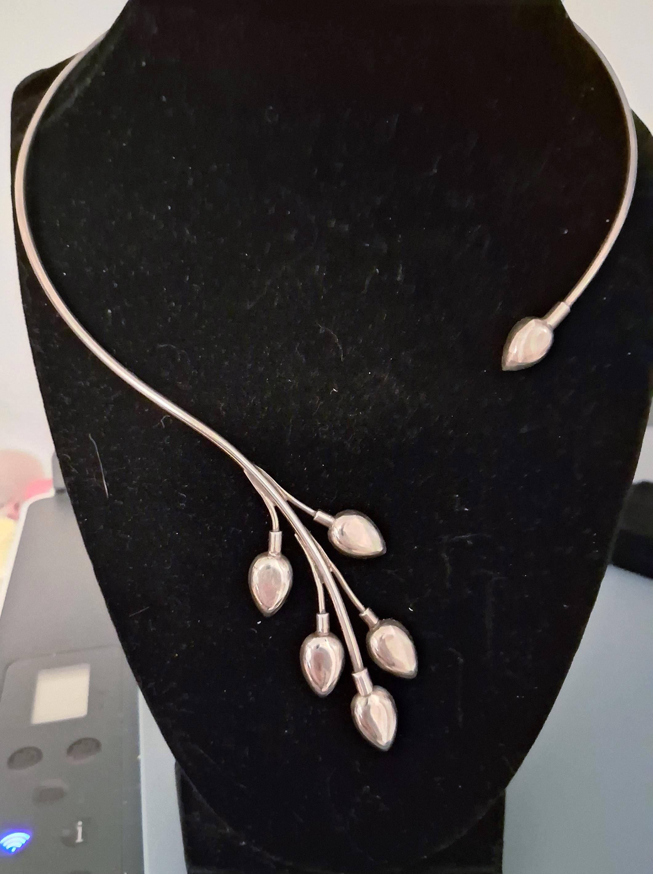 May include: A silver-toned metal collar necklace featuring a curved, open-ended design. One end terminates in a single teardrop-shaped charm, while the other extends into a branch-like structure adorned with five matching teardrop charms.