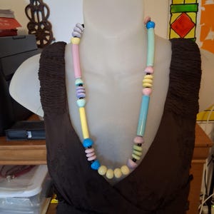 May include: A long necklace featuring a variety of smooth, cylindrical and disc-shaped beads in pastel shades of pink, yellow, light blue, lavender, and mint green, interspersed with small black spacer beads on a dark string.