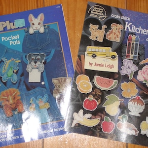 May include: Two cross stitch pattern books. The first book is blue and features a variety of pocket pals, including a teddy bear, a dragon, a frog, a dinosaur, a cat, and a dog. The second book is purple and features a variety of kitchen magnets, including a cat, a dog, a school bus, crayons, a carrot, a flower, a watermelon, a pear, an apple, a banana, a sundae, and an ice cream cone. The text on the second book reads "Kitchen Magnets on Perforated Paper" and "by Jamie Leigh".