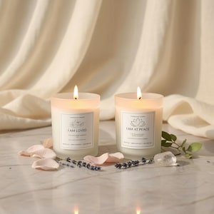 May include: Two lit, frosted glass candles with labels that read "I AM LOVED" and "I AM AT PEACE." The candles are surrounded by rose petals, lavender sprigs, and a clear crystal on a marble surface, with a cream-colored fabric backdrop.