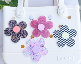 Könnte beinhalten: Weiße Tragetasche mit fünf blütenförmigen Anhängern verziert. Die Anhänger weisen verschiedene Muster und Farben auf, darunter Leopardenmuster, glitzerndes Rosa, Schachbrettmuster und Pastelltöne. Die Tasche hat einen Griff und die Marke "bogg" ist sichtbar.