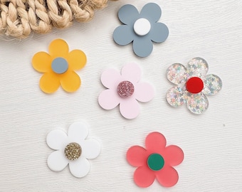 Puede incluir: Seis adornos planos de acrílico con forma de flor en varios colores y acabados. El conjunto incluye una flor gris con centro blanco, una flor amarilla con centro gris, una flor rosa claro con centro brillante, una flor transparente con purpurina y centro rojo, una flor blanca con centro de purpurina dorada y una flor coral con centro verde.