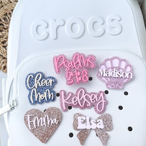May include: A collection of glittery charms in various shapes and colors, including a heart, bow, and seashell, with names and phrases like "Cheer Mom," "Psalms 24:8," and "Elsa." These charms are designed to be attached to a white backpack.