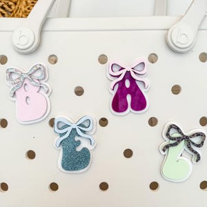 May include: Close-up of a white tote bag with several decorative letter charms. The charms are in the shape of the letters A and B, with bow accents. The letters are in various colors, including pink, teal, and green.