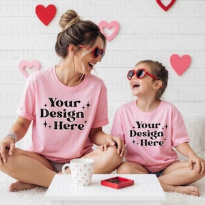 May include: Two pink t-shirts with the text "Your Design Here" are worn by a woman and a child. Both are wearing heart-shaped sunglasses. A white table holds a mug and a small red box. Decorative hearts are on the wall.