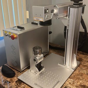 May include: A silver laser engraving machine with a rectangular base and a tall, vertical support. The machine has a control panel with buttons and a red emergency stop button. A black case sits to the left of the machine.