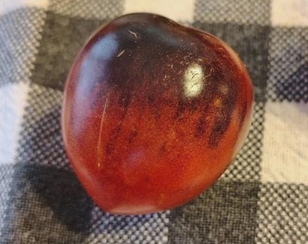 May include: A ripe, heirloom tomato with a deep red and purple gradient. The tomato is oval-shaped and rests on a black and white chequered surface. The skin has a smooth, glossy texture.