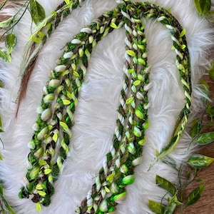 May include: A set of long, braided hair extensions featuring a blend of brown, white, and various shades of green yarn. The braids are interwoven with artificial moss and small, bright green fabric leaves, creating a textured, multi-strand accessory.