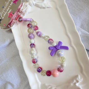 May include: A clear phone case with a pink and gold star design and the text "Augenstern the stars don't fall for men." A beaded phone charm features pink, purple, and clear beads, a purple bow, and a pink bead.