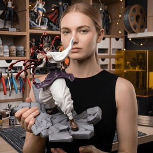 Puede incluir: Figura detallada de un personaje con un atuendo blanco y morado con detalles en rojo, de pie sobre una base rocosa gris. La figura la sostiene una persona con una parte superior negra. Estantes en el fondo muestran otras figuras y materiales de arte.