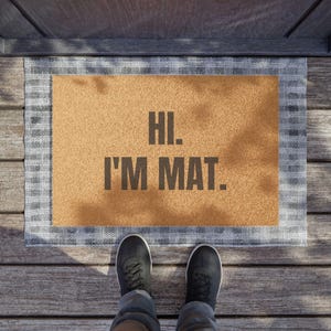 Puede incluir: Felpudo rectangular marrón con el texto "HI. I'M MAT." en letras grises oscuras. El felpudo tiene un borde gris y blanco estampado. Se ven zapatos negros y vaqueros azules.