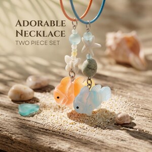 Op de afbeelding: Tweedelige ketting set met oranje en blauwe vishangers, versierd met kralen, zeesterren en vlinderbedels. De kettingen hangen aan oranje en blauwe koorden. De tekst luidt: "ADORABLE NECKLACE TWO PIECE SET".