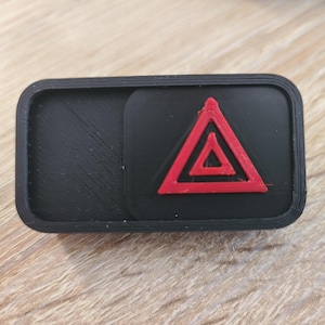May include: A black rectangular button with a red hazard symbol. The hazard symbol is a red triangle with an inner triangle. The button has a textured surface.