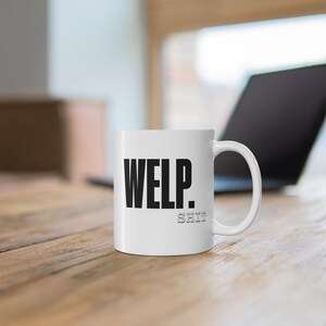 Puede incluir: Taza de cerámica blanca con la palabra "WELP." en letras negras en negrita y la palabra "SHIT" en letras negras más pequeñas y delineadas. La taza está sobre una superficie de madera, con un portátil en el fondo.