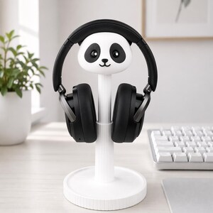 May include: A white panda-shaped headphone stand holding black headphones. The stand has a round base and a vertical support with a panda face at the top. The headphones are black with silver accents.