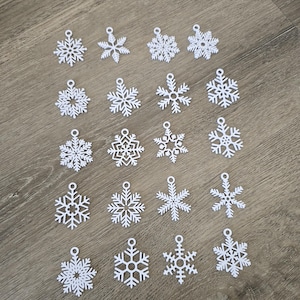 Op de afbeelding: Een verzameling van twintig witte sneeuwvlokornamenten, elk met een uniek ontwerp. De ornamenten zijn gemaakt van een lichtgewicht materiaal en hebben een kleine lus aan de bovenkant om op te hangen. Ze zijn gerangschikt op een houten oppervlak.
