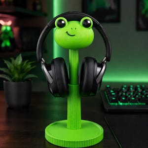 May include: A vibrant green frog-shaped headphone stand. The stand has a smiling frog face, a vertical stem, and a circular base. Black headphones are placed on the stand. The stand is on a dark surface.