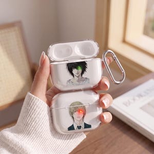 May include: A clear plastic protective case for wireless earbuds featuring two anime-style character illustrations. The top lid shows a dark-haired character with a glowing green eye, while the bottom section displays a blonde character with a glowing red eye.