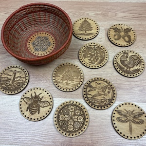 May include: A woven basket with a red and brown design, accompanied by a set of wooden coasters. The coasters feature various engraved designs, including a tree, grapes, a rooster, and a dragonfly. The coasters are round and have a decorative border.