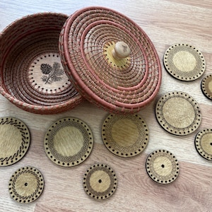 May include: A woven basket with a lid and a set of wooden coasters. The basket is brown and pink, with a wooden knob on the lid. The coasters are round, with decorative edges and a central hole.