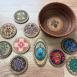 May include: A collection of nine circular and one oval wooden base pieces featuring various colorful printed designs, including geometric patterns, floral motifs, a sea turtle, and a stained glass style, alongside a round woven basket with a pinecone.
