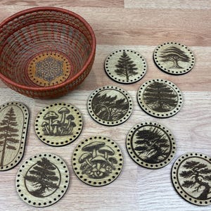 May include: A woven basket with a red rim and a wooden base, alongside a collection of wooden coasters. The coasters feature laser-cut designs of trees and mushrooms. The coasters are round and oval, with a light wood color.