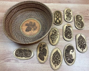 Puede incluir: Una cesta tejida con un diseño de piña en la base, rodeada de varias piezas ovaladas de madera. Cada pieza presenta un diseño grabado diferente inspirado en la naturaleza, incluyendo árboles, flores y hojas. La cesta y las piezas están sobre una superficie de madera clara.