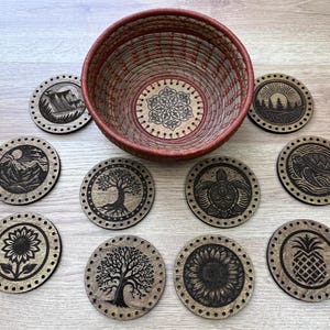 May include: A collection of wooden coasters with various nature-themed designs, including mountains, trees, a turtle, and a sunflower. A woven basket with a floral design sits in the center. The coasters are arranged on a light wood surface.