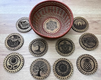 Puede incluir: Un conjunto de posavasos de madera con diseños grabados de montañas, árboles y una tortuga, junto con una cesta tejida. Los posavasos se exhiben sobre una superficie de madera clara. Cada posavasos tiene un diámetro de aproximadamente 10 cm.