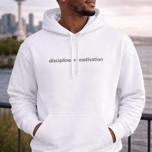 May include: White hoodie with the text "discipline > motivation". The hoodie features a kangaroo pocket and a drawstring hood. The background shows a cityscape and water.