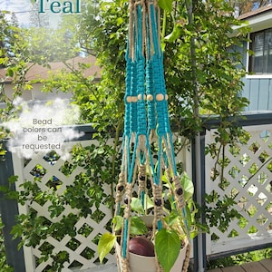 May include: A teal and beige macrame plant hanger with wooden beads and a white pot holding a plant. The text "Mystic Teal" and "Bead colors can be requested" are visible. The plant hanger is suspended outdoors.