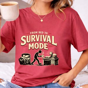 May include: Red t-shirt with the text "FROM BED TO SURVIVAL MODE" in a vintage style. The graphic depicts a stick figure walking from a bed to a desk with a skull, books, and a lamp. The person is holding a coffee cup.