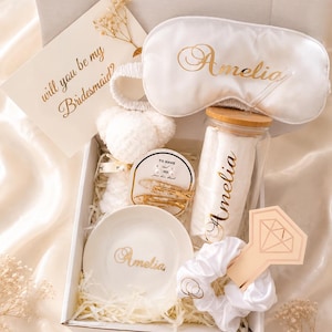 Könnte beinhalten: Eine weiße Geschenkbox mit einer weißen Schlafmaske mit dem Namen Amelia in Gold, einem Glasbecher mit Bambusdeckel und dem Namen Amelia, einer weißen Keramikschale mit dem Namen Amelia, einem weißen Haargummi, zwei Haarspangen und einer Karte mit der Aufschrift will you be my Bridesmaid?